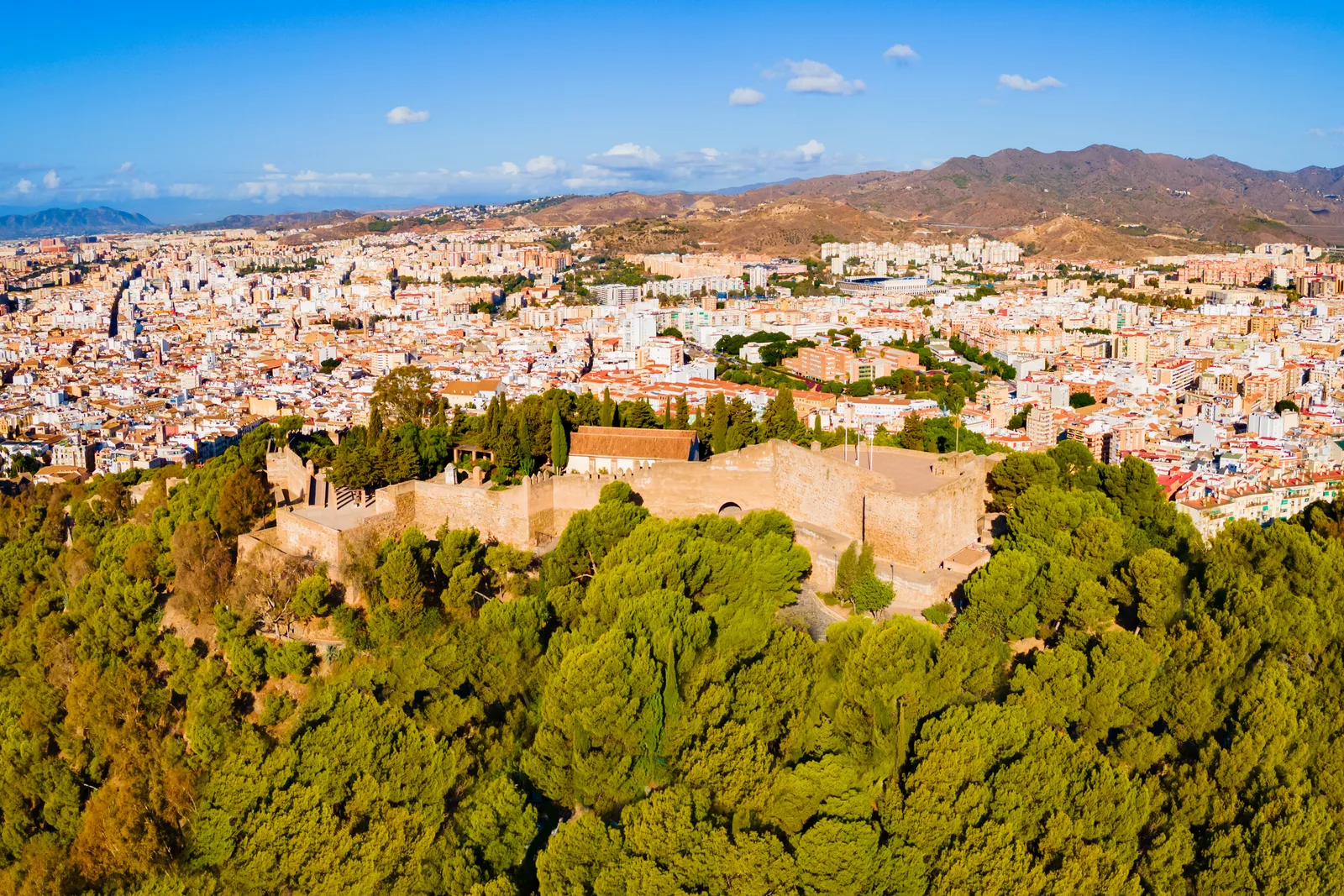 Vista aérea de las murallas del castillo entre pinos en lo alto del monte, con la ciudad y las sierras detrás