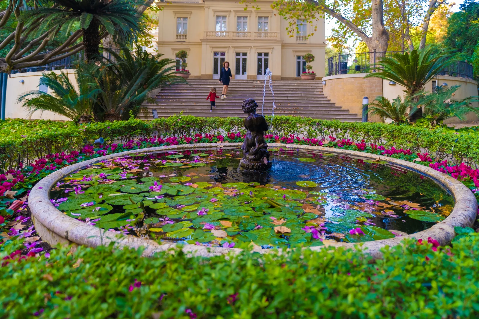 Fuente redonda con nenúfares y flores ante la escalinata de la casa-palacio del jardín, rodeada de palmeras