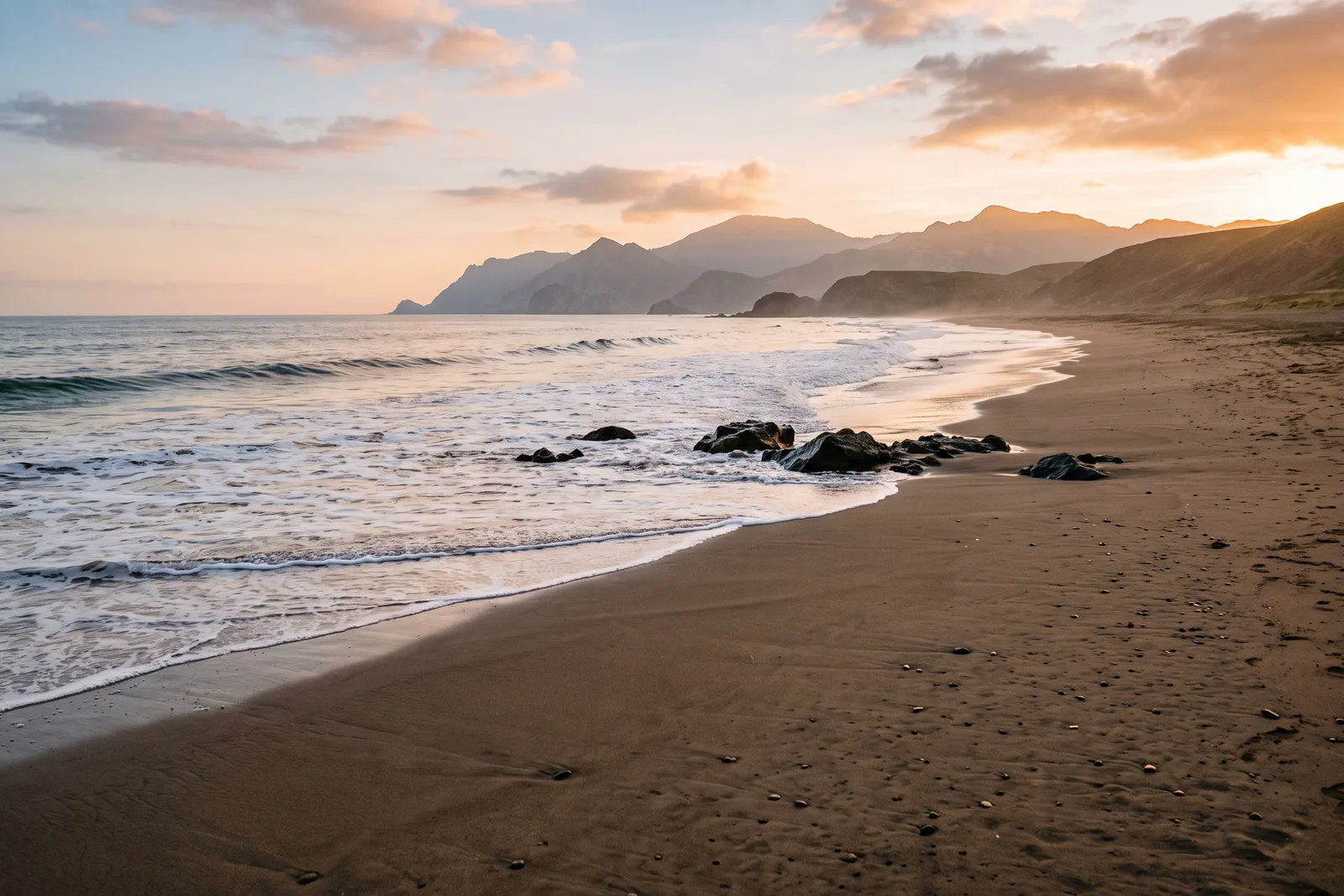 Orilla de arena con rocas oscuras y montañas al fondo bajo un cielo de atardecer