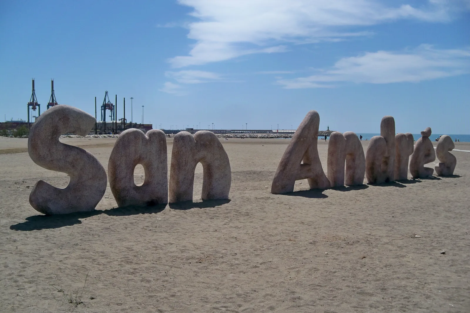 Letras gigantes con el nombre de la playa sobre la arena y las grúas del puerto al fondo