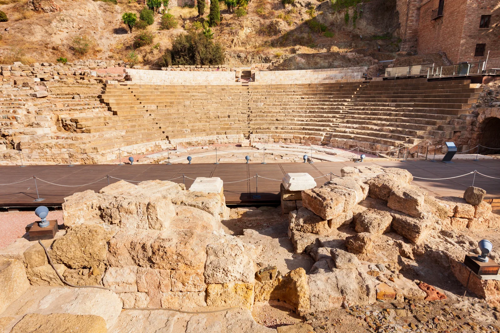 Gradas semicirculares de piedra del teatro romano con restos de sillares en primer plano y la ladera de la Alcazaba encima